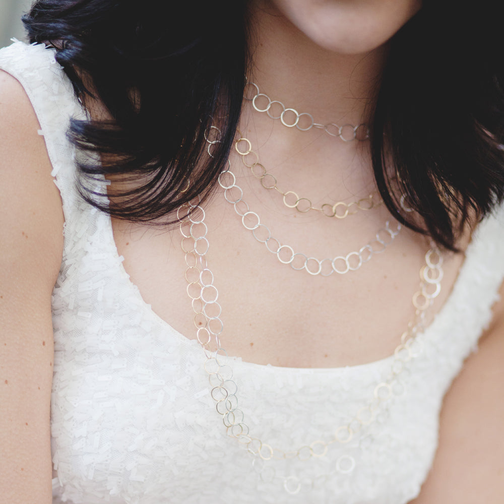 Multiple hammered chains in sterling silver and gold-filled on a white woman wearing a white sequin dress.